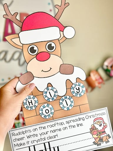 Reindeer wearing a Santa hat above a rooftop with letter snowflakes for name practice